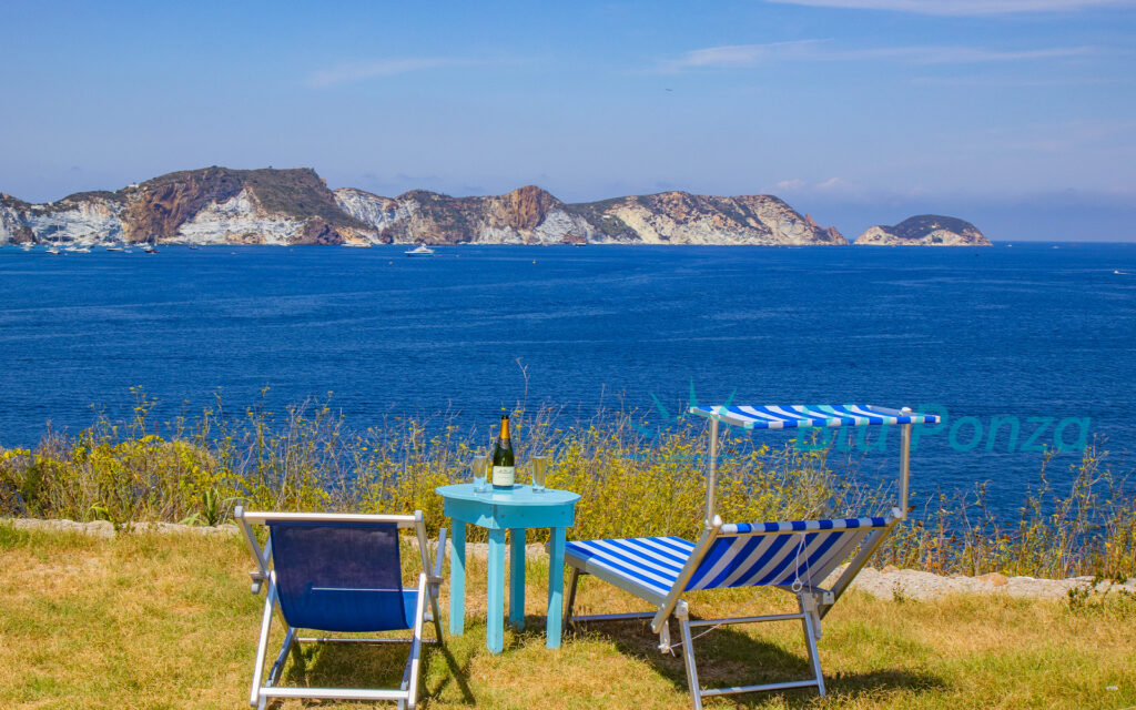 Vista panoramica sul mare a Ponza con due sedie a sdraio e un tavolino azzurro con una bottiglia di champagne, in un giardino affacciato sull'isola di Ponza.